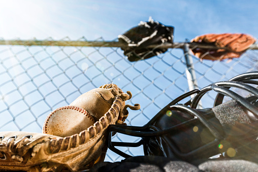 Looking up at a close-up of a  baseball in a brown leather catcher's mitt along with a catcher's mask and chest protector sitting on an aluminum bench with a chainlink backstop and blue sky in the background along with lens flare from the sunshine