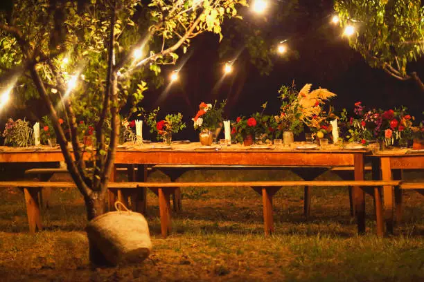 Prepared table for a rustic outdoor dinner at night with wineglasses, flowers and lamps in the garden