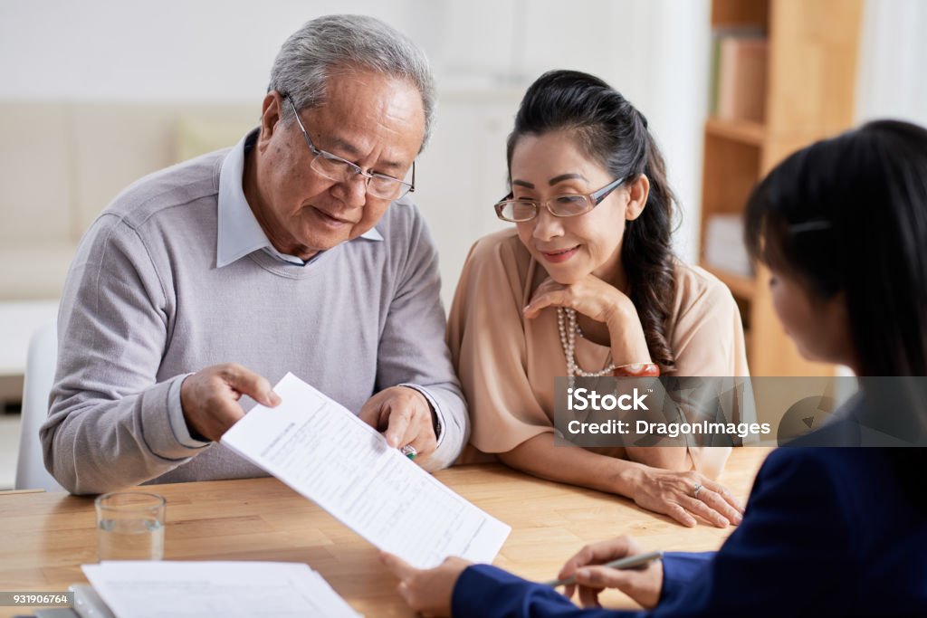 Studying Real Estate Purchase Agreement Concentrated senior man and his pretty wife studying terms of real estate purchase agreement while having meeting with Real Estate Agent, interior of modern office on background Planning Stock Photo