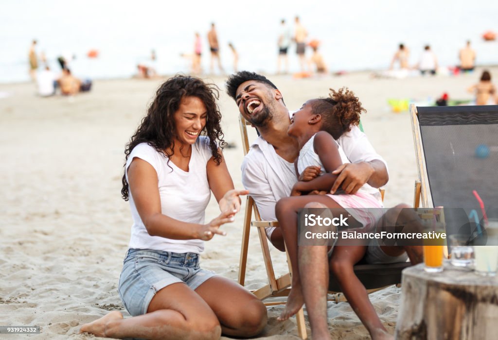 family on beach Young mixed race family sitting and relaxing at the beach on beautiful summer day.Daughter lies in father lap and laughing. Family Stock Photo