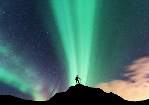 Aurora and silhouette of standing woman on the top of mountain. Lofoten islands, Norway. Aurora borealis and happy girl. Sky with stars and green polar lights. Night landscape with aurora and traveler