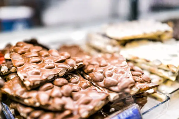 Macro closeup of pile of many milk dark brown chocolate bar almond nut peanut pieces chunks on tray glass display in candy store chocolatier shop