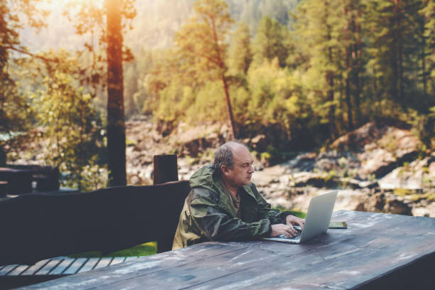 Businessman with netbook during his vacations Elderly man employer is spending his vacations with work outdoors: sitting with the laptop at the wooden table with a digital tablet next to him and summer pine forest in a defocused background woodward stock pictures, royalty-free photos & images