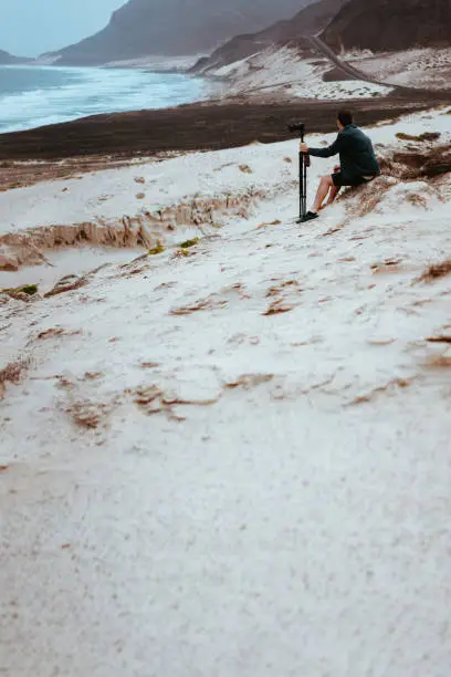 Photo of Photographer sitting and admitting bizarre landscape of sand dunes volcanic cliffs on the Atlantic coast. Baia Das Gatas, near Calhau, Sao Vicente Island Cape Verde