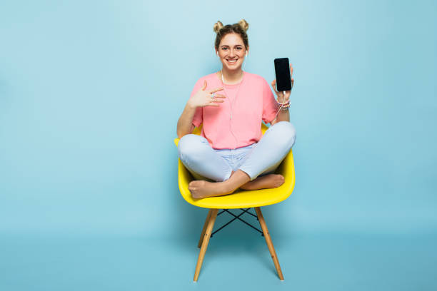 woman sitting on a chair with crossed legs and points to her smartphone modern young woman sitting on a chair with crossed legs and points to her smartphone against a blue pastel background . youth lifestyle girl sitting stock pictures, royalty-free photos & images