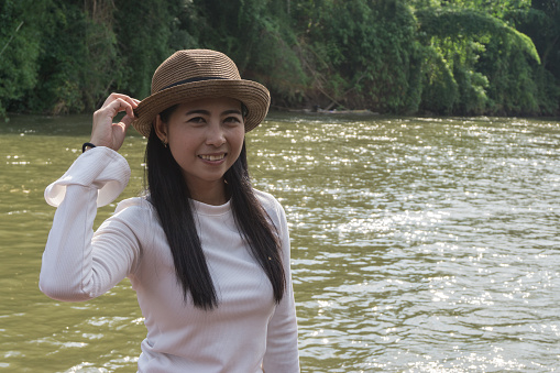 Happy Asian girl white shirt and brown hat on the river in nature background, Relax time on holiday concept travel., Thailand river kwai and typical landscape kanchanaburi., with copy space for text