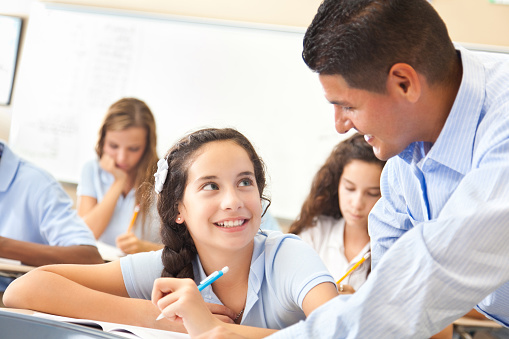 A smiling preteen girl sits at a desk during class at school.  She looks up and listens as her male teacher leans over and praises her hard work.