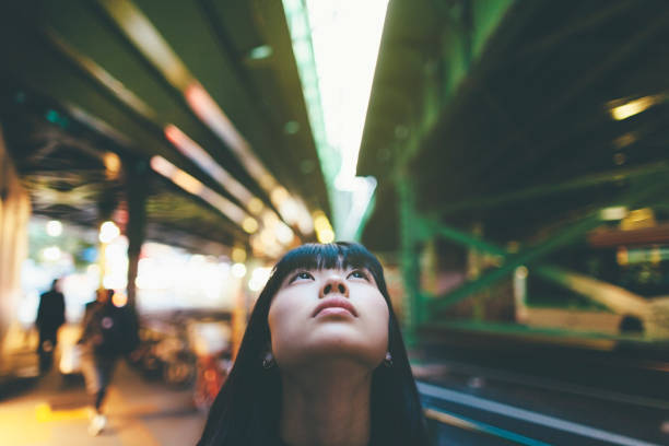 Close up portrait of woman while loo0king up in the city A young woman is looking up between two bridges in the city. japanese woman stock pictures, royalty-free photos & images