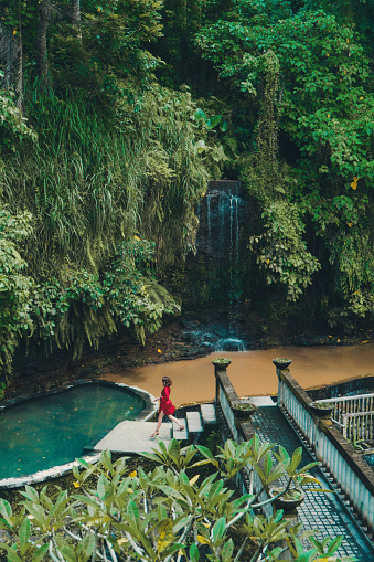 Young Caucasian woman  in red bathrobe walking near the swimming pool and waterfall in Bali