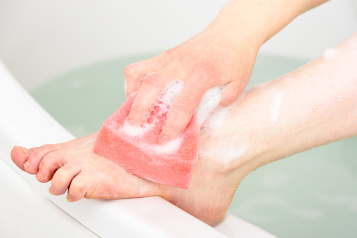 Caucasian woman washing her leg and foot in a bathtub using a pink bathing sponge.