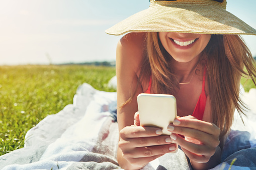 Shot of a cheerful young woman wearing a hat while lying on the ground busy with her phone outside in the sun