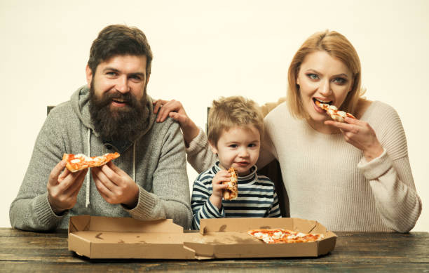 happy family eating pizza on the wooden table. family takes pieces of pizza. happy lovely family eating pizza. young mother and her little son eating pizza and having fun with husband. family kitchen - two parent family indoors home interior domestic kitchen imagens e fotografias de stock