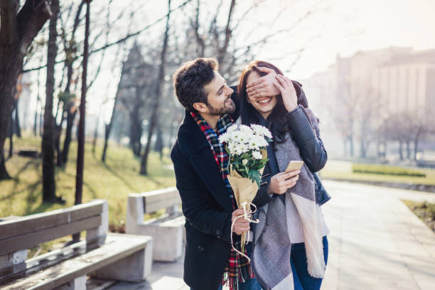 Young couple dating on one sunny spring day Cheerful young man surprising his girlfriend with spring flowers bouquet and hiding her eyes with hand gift for girlfriend stock pictures, royalty-free photos & images