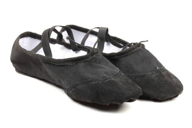 Young adult female ballerina with brunette hair and red flower accessory sits on studio floor. Dressed in black attire and pointe shoes, she exudes elegance. Soft lighting enhances serene atmosphere