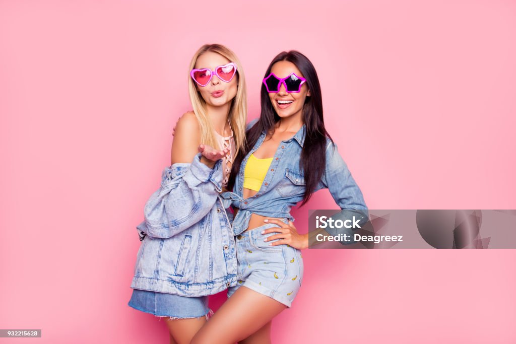 I like you! Beautiful playful cheerful women dressed in fashionable stylish shorts, shirt, jacket, top, funny star and heart glasses are embracing, sending air-kiss, isolated on bright pink background Denim Stock Photo