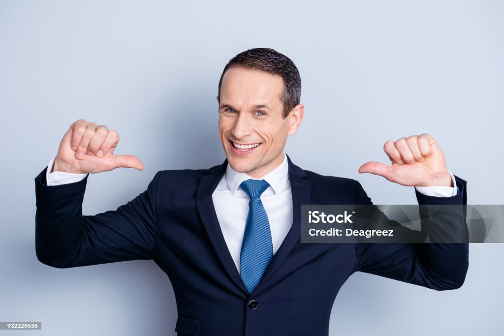 Cheerful financier, positive economist, confident politic man in formalwear with tie showing with two thumbs on himself,  standing over gray background Pointing Stock Photo