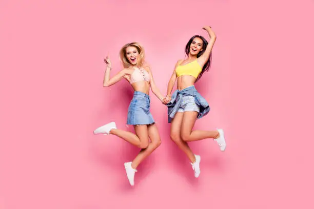 Photo of Life is cool! Beautiful attractive funny joyful cheerful relaxed carefree girls clothed in casual trendy outfit and white shoes are jumping up ans holding hands, isolated on bright pink background