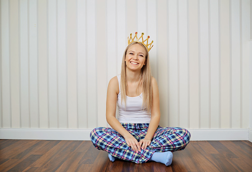 Funny blonde girl in pajamas with crown on her head smiling sits on the floor against a striped wall background. Young woman princess laughs in the room.