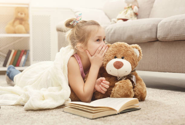 happy little female child and her teddy bear reading book on the floor at home - segredo criança imagens e fotografias de stock