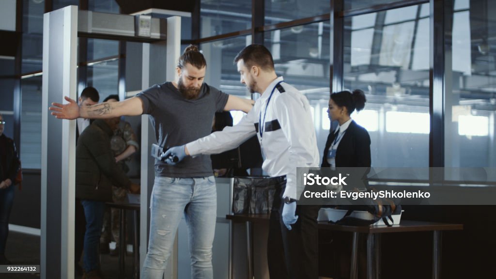 Security agent patting down a male passenger Security agent at an airport check-in gate patting down a bearded casual male passenger with outstretched arms after he passes through the metal detector scanner in the departures hall. Airport Stock Photo
