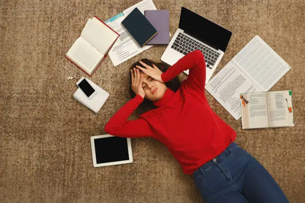 Exhausted student girl lying on the floor among textbooks, tests and gadgets, copy space. Woman holding head with hands, got tired while preparing for exams. Education and overworking concept