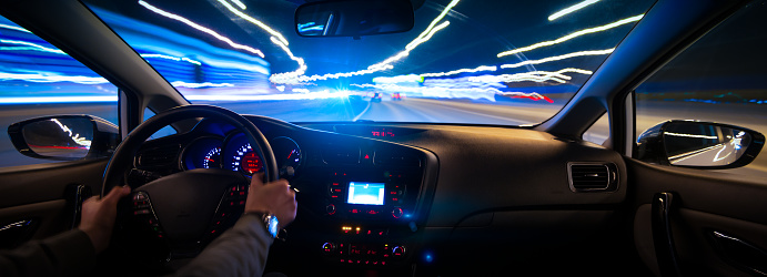 Hands on the wheel the car moves with fast speed on the highway at night. Blurred road with lights with a car at high speed. High beam headlights blinding