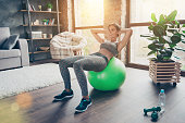 Pretty beautiful powerful skinny cheerful woman clothed in sportive outfit lying on green fit ball and doing crunches at home in sun beam, light, glare, sunny day, reflection