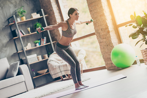 Low-angle photo of sportive strong active attractive purposeful ideal woman with excellent body figure doing exercise with lifting green dumbbells at home in front of tv in sun glare, sunlight
