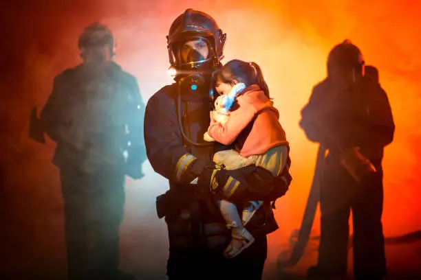 The team of firefighters dressed in protective suits with protective fire helmets. The firefighter is holding the little girl on his hands on foreground. He is seriously looking at the camera. Other firemen are holding the equipment firefighter gear in their hands and standing behind. Studio shooting with a red lighting and smoke