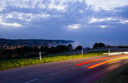 Cars driving on the asphalt country road over the sea bay at twilight, Normandy, France. Countryside landscape with dramatic sky. Road network and transportation concept