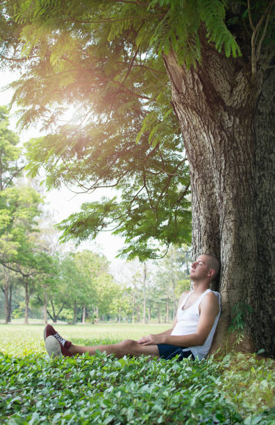 Male runner resting against tree in park with eyes closed.