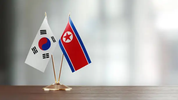 South Korean and North Korean flag pair on desk over defocused background. Horizontal composition with copy space and selective focus.