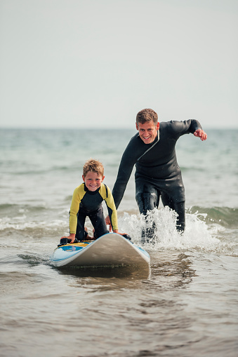 Little boy getting shown how to surf at the beach with his father.