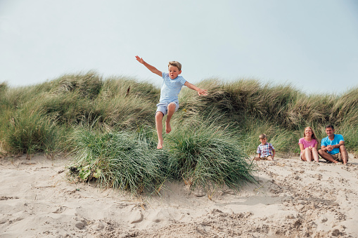 Wide angle shot of a little boy having fun while jumping over a sand dune. His family wait, watching at the top of the dune.