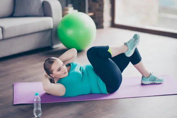 Photo of Cheerful active excited concentrated positive model fatty woman is improving muscles on her stomach, she is doing crunches on the floor at home