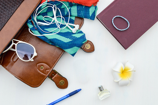 Contents of a brown leather purse consisting of a pen, sunglasses, scarf, earbuds, hair tie, book, flower and perfume laid out on a white table.