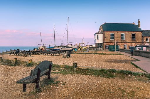 Whitstable, Kent, UK beach with wooden benches and old wooden boats moored on the beach. There is a red brick building and a pedestrian pathway. There equipment machinery of the oyster industry.