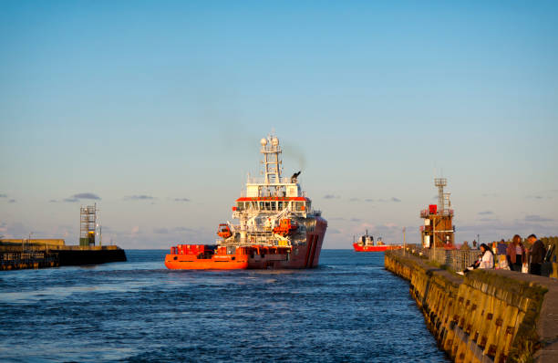 People enjoying the open late summer air as two rig support vessels, including the “Forties Sentinel”, leaving the River Yare between Gorleston-on-Sea and Great Yarmouth, Norfolk, Eastern England, in warm evening light. The “Forties Sentinel” belongs to Sentinel Marine and is classed as a ‘Multi-Role Field Support Emergency Response and Rescue Vessel’. Other people are fishing from the jetty in Gorleston-on-Sea.