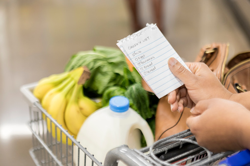 Female grocery store customer checks items off of her grocery list. Banana, milk and leafy green vegetables are in the grocery cart.