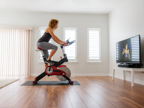 Woman Exercising on Spin Bike in Home A woman exercising on a spin bike using an online instructor inside a home. exercise bike stock pictures, royalty-free photos & images