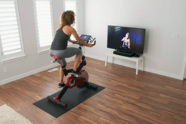 Woman Exercising on Spin Bike in Home A woman exercising on a spin bike using an online instructor inside a home. exercise bike stock pictures, royalty-free photos & images