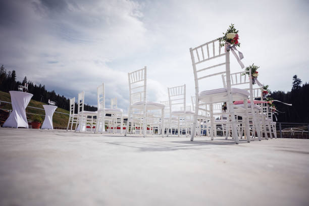 A tranquil and elegant outdoor scene captures a neat stack of white chairs placed on a minimalist green lawn in a garden setting. The area is prepared for an outdoor celebration, subtly enhanced by decorative string lights suspended among the surrounding trees. This visual, often associated with white wedding chairs, powerfully conveys themes of serenity, organized anticipation, and the simple elegance of a romantic event held in a beautiful, natural setting.