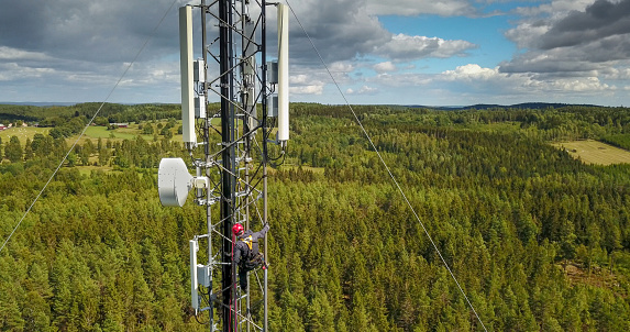 Telecommunication manual high worker engineer installing new 4g, 5g LTE antenna on tall mobile base station (communication tower) in the middle of european forest. Working at height. Telecommunication masts and towers are typically tall structures designed to support antennas for telecommunications and broadcasting. Drone point of view.