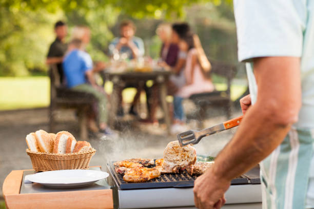 Preparing food at picnic Rosting meat on barbecue for a family picnic in the back. Sun through the trees. Family at the table. grilled stock pictures, royalty-free photos & images
