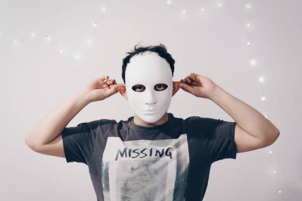 istock Portrait Of Man Wearing Mask While Holding Ears Against String Lights Decorated White Wall 932506836