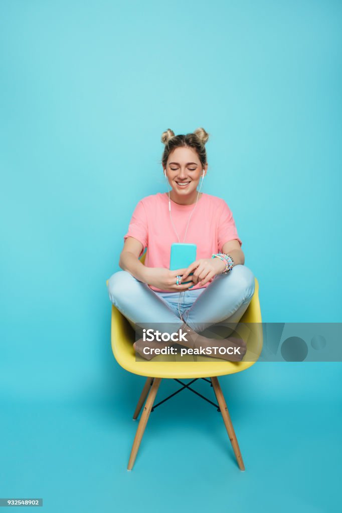 woman sitting on a chair with crossed legs and listening to music enjoy the music. modern young woman wearing headphones listening to music, sitting on a chair with crossed legs against a blue pastel background Women Stock Photo