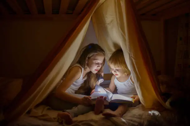 Photo of Two girls read fairy tales at dark night under a blanket by the light of a small lamp