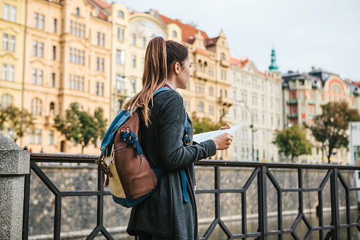 A young beautiful girl stands and looks at the map next to the Vltava River with the amazing old architecture of Prague in the background with a sunny spring day. Interesting attraction. Tourism