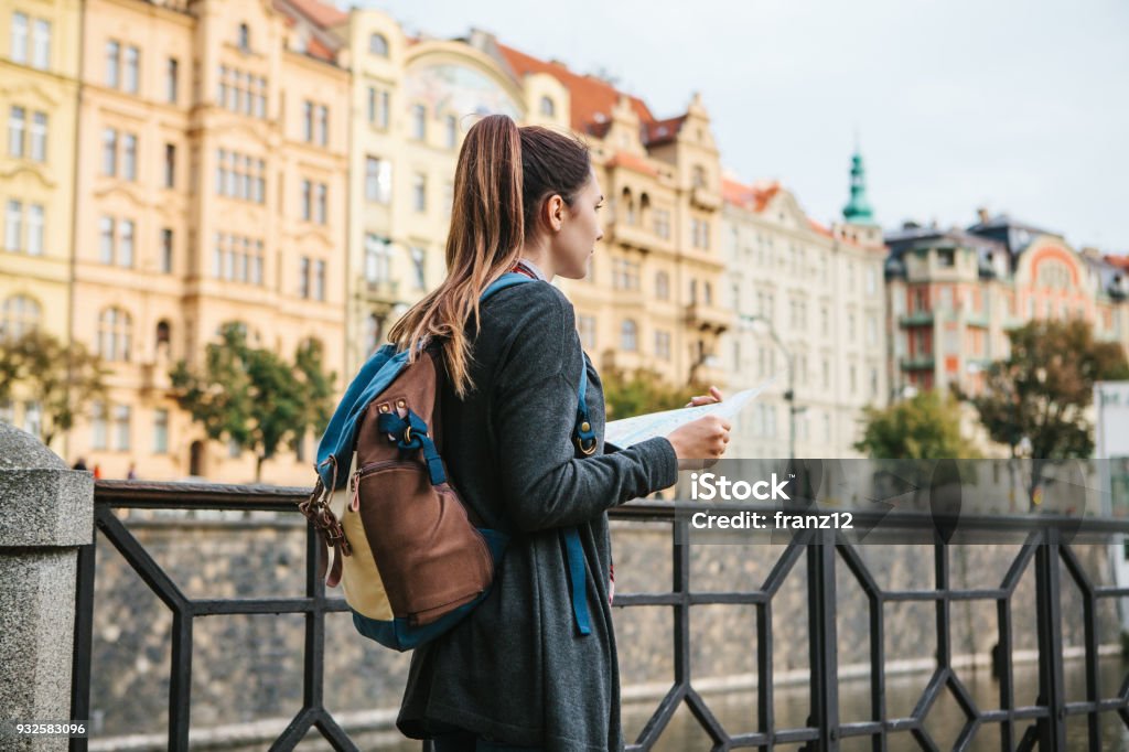 Uma bela jovem representa e olha para o mapa ao lado do Rio Vltava, com a incrível arquitetura antiga de Praga em segundo plano com um dia ensolarado de primavera - Foto de stock de Turista royalty-free