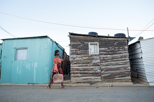 A black woman in colourful clothes walks past the shacks on her way home from work, Kayamandi - Cape Town